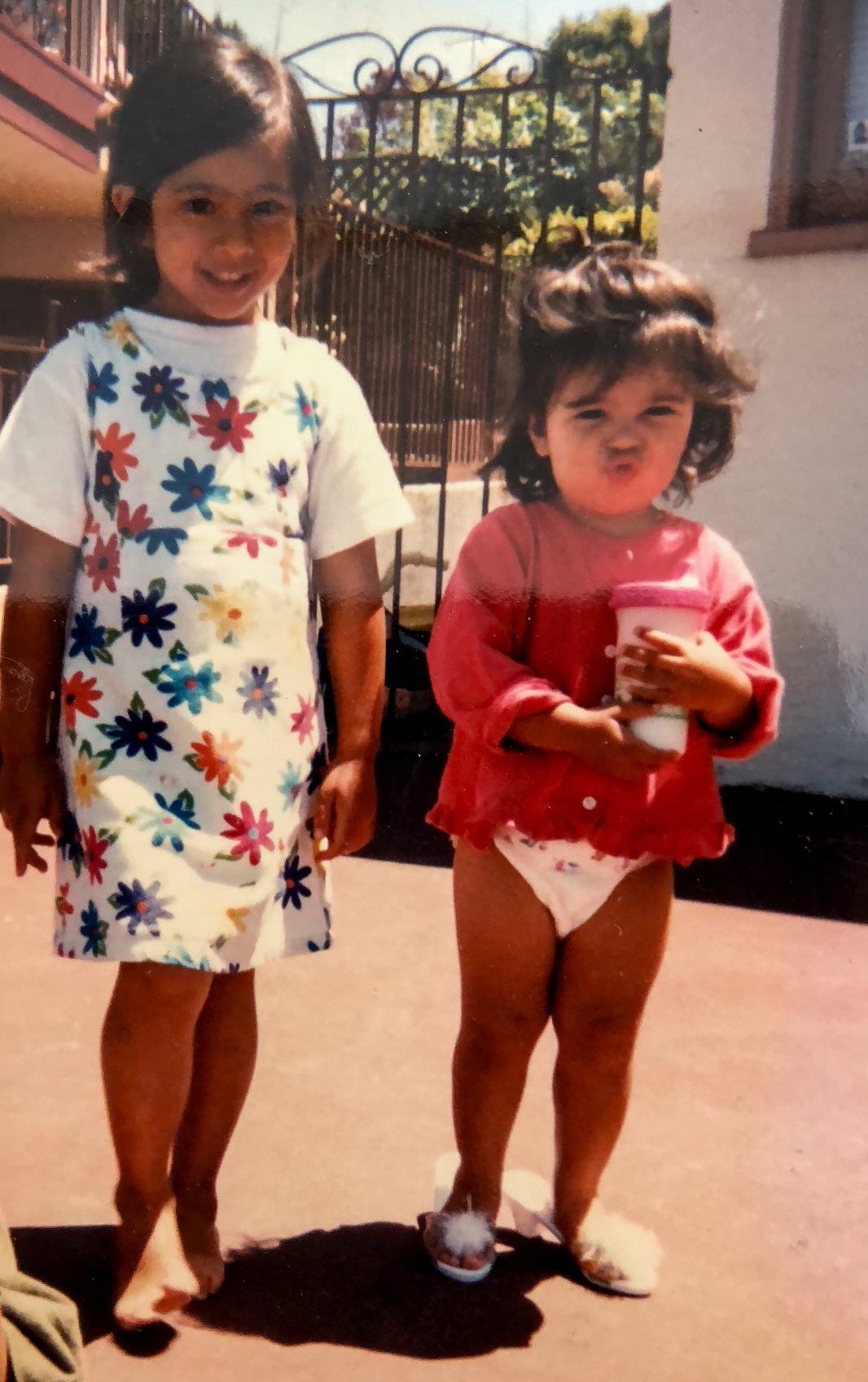 Gemma as a toddler in a red top and diaper, pouting and holding a cup, next to her sister in a flowered dress