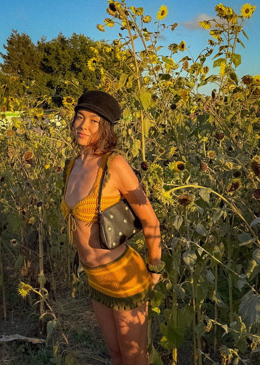Woman in a mustard crochet bikini set and cap standing in a sunflower field at golden hour, with a small studded bag
