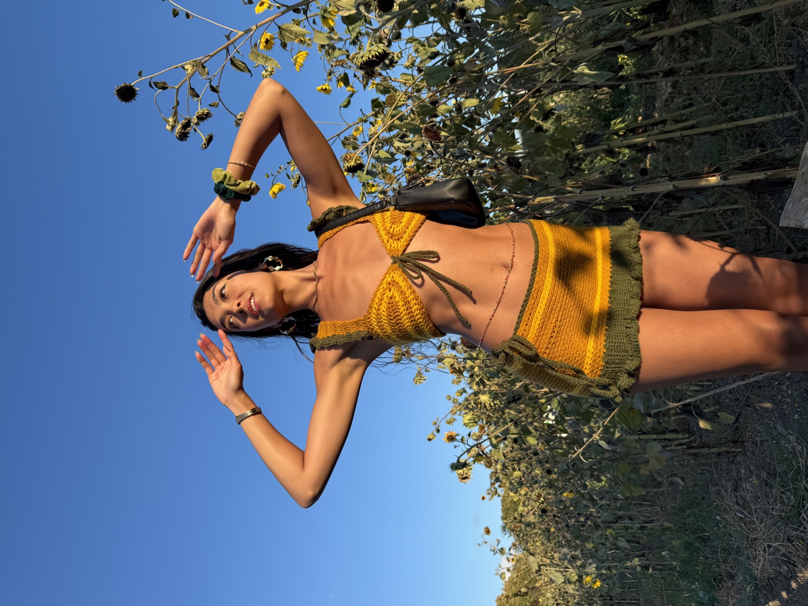 Woman in the mustard crochet set with arms raised among tall sunflowers