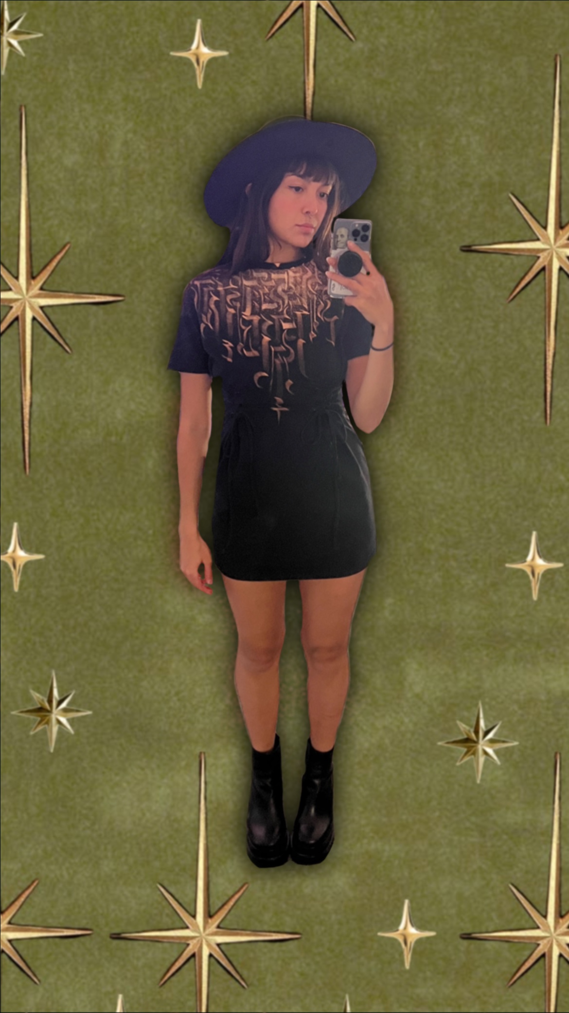 Mirror selfie in a black graphic-print dress with a wide-brim hat, over a starry green backdrop
