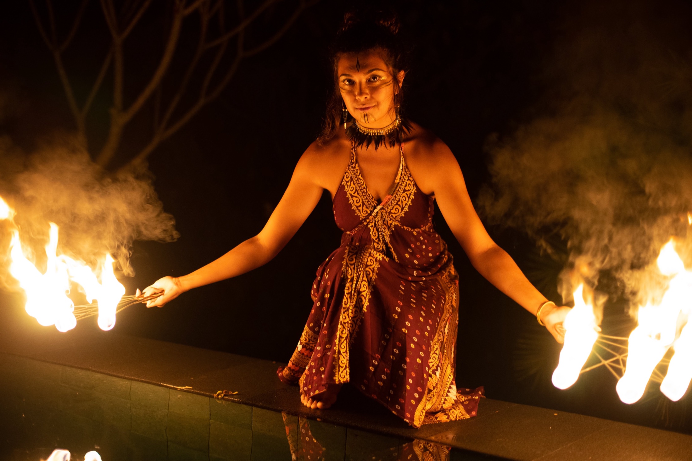 Gemma kneeling with tribal makeup and a batik dress, holding two fire fans alight on either side