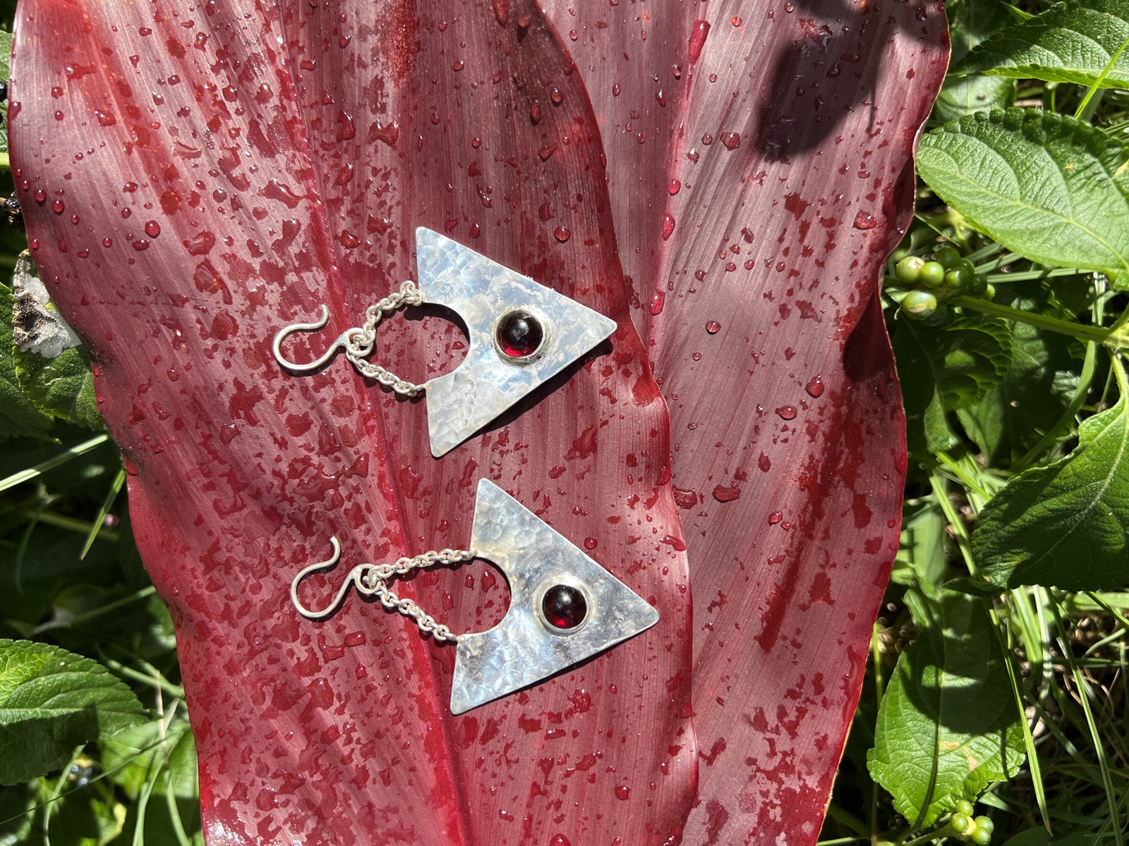 A pair of hammered silver triangle earrings set with garnet, laid on a rain-speckled red ti leaf