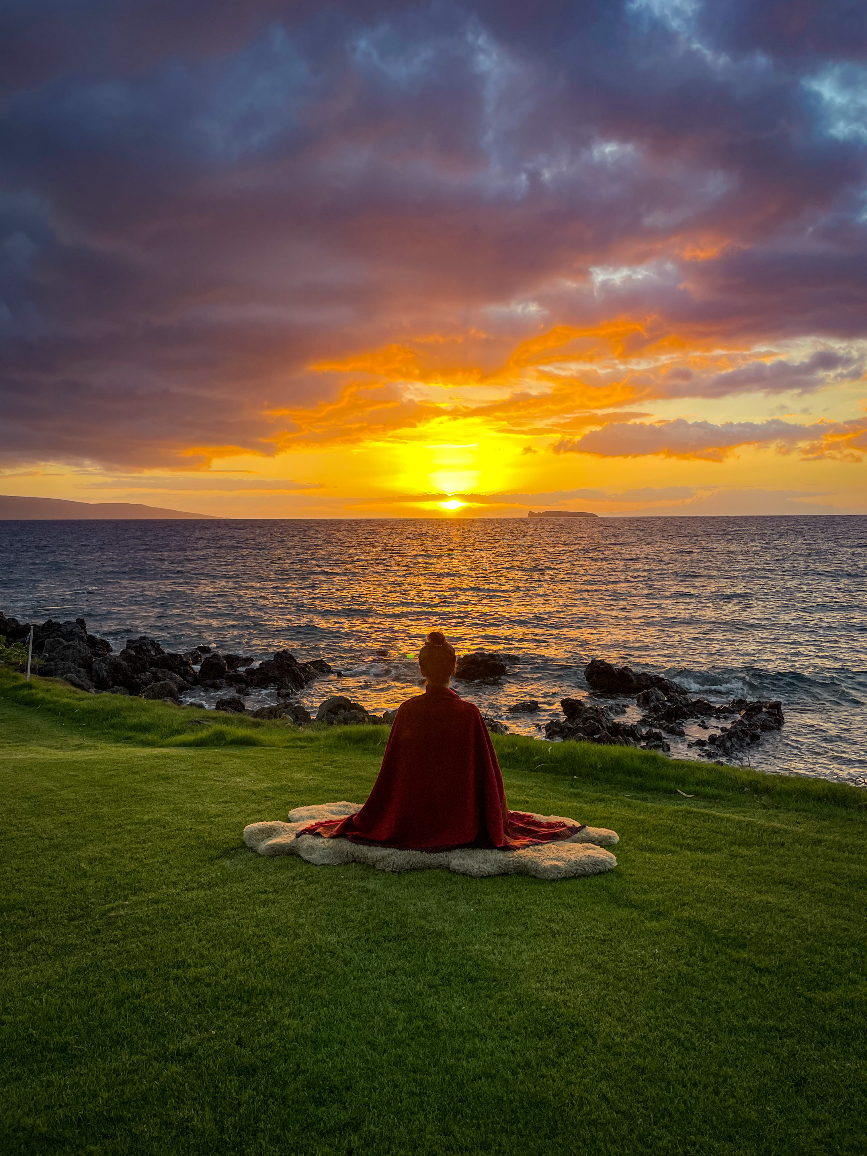 Gemma Rose meditating on the grass overlooking the ocean at sunset in Maui