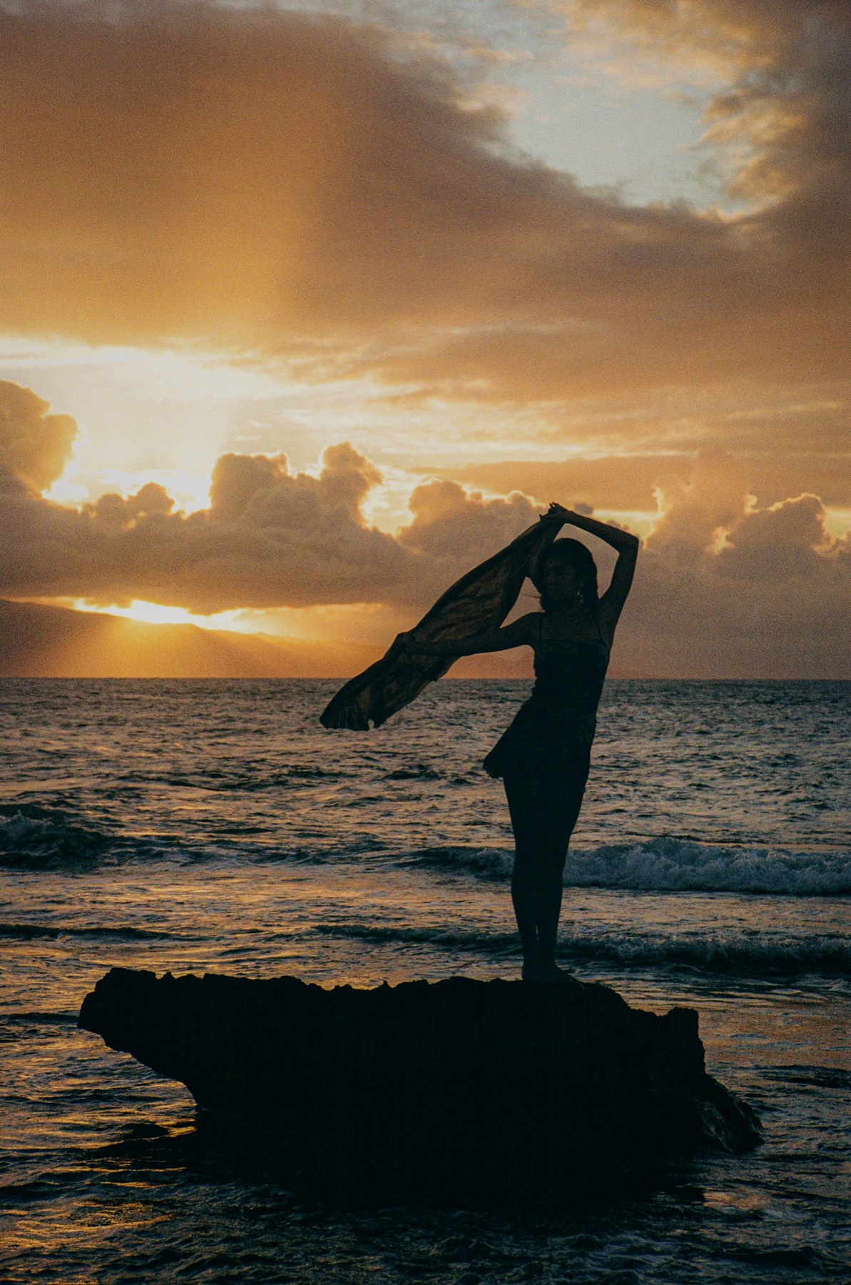 Gemma Rose in silhouette on a rock at sunset, holding a scarf billowing overhead
